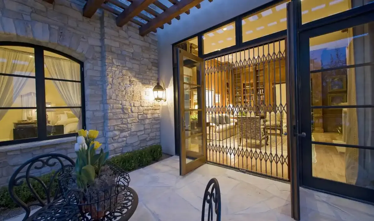 A dark trellis gate locked across a courtyard doorway at dusk, with the lit room behind it visible through the flights.